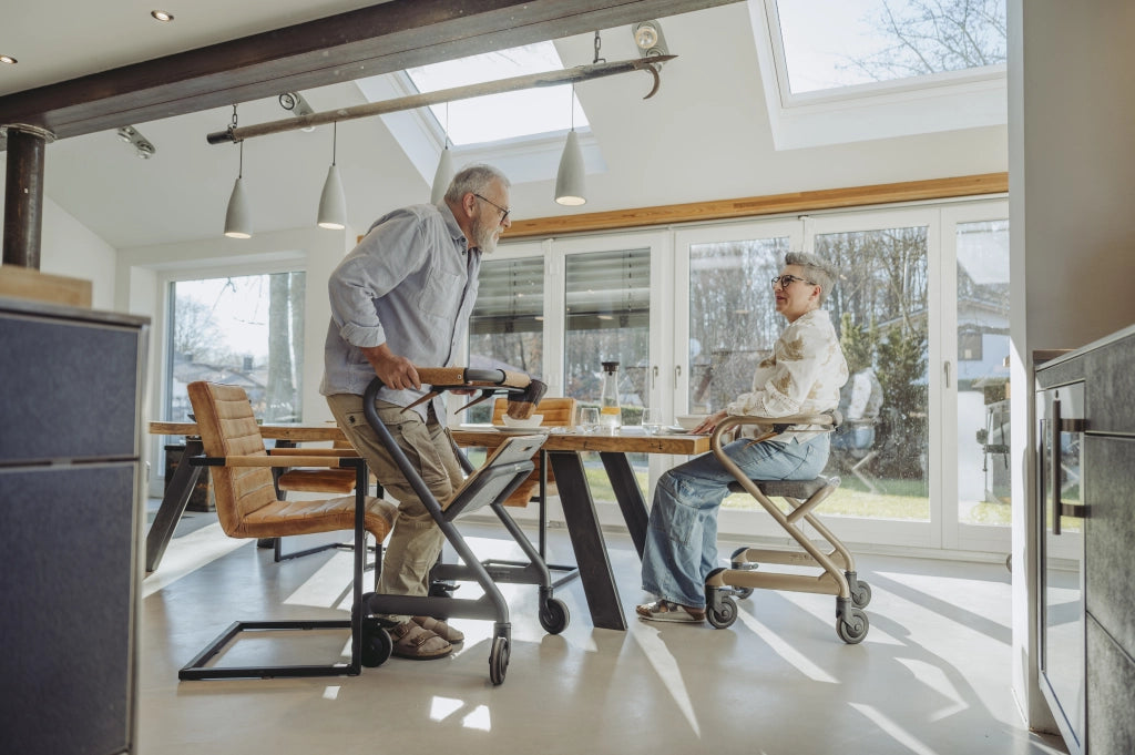 Seniorenpaar beim Frühstück am Esstisch mit SALJOL Wohnraumrollatoren WR 2025 – Mann steht am Rollator in Velvet Grey, Frau sitzt entspannt auf Rollator in Champagne Gold, lichtdurchfluteter Raum mit großen Fenstern