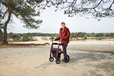Ältere Frau mit rotem Mantel nutzt den Rollz Motion Performance Rollator in Ruby Red bei einem Spaziergang durch eine Dünenlandschaft.
