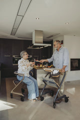 Seniorenpaar in moderner Küche mit SALJOL Wohnraumrollatoren WR 2025 – Frau sitzt entspannt auf dem Rollator, Mann serviert Speisen über das Tablett, beide Modelle in unterschiedlichen Farben