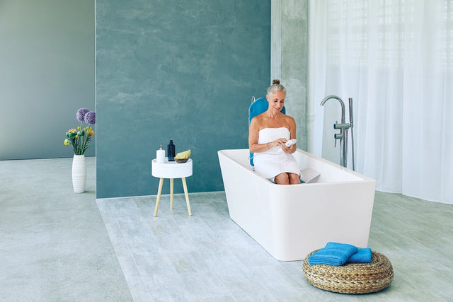 Eine ältere Frau sitzt entspannt in einer freistehenden Badewanne auf einem Badewannenlift mit blauer Rückenlehne und nutzt eine Fernbedienung. Das stilvolle Badezimmer ist modern eingerichtet.